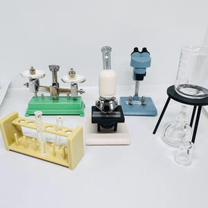Op de afbeelding: Een set miniatuur wetenschappelijke apparatuur, waaronder een weegschaal, een microscoop, een reageerbuisrek, bekers en een standaard. De apparatuur is gemaakt van plastic en metaal en is geverfd in verschillende kleuren, waaronder groen, blauw en zwart.