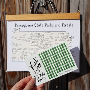 May include: A white card with black text that reads "Visit all 124 PA State Parks" and a green pattern of trees. The card is held in front of a map of Pennsylvania State Parks and Forests.