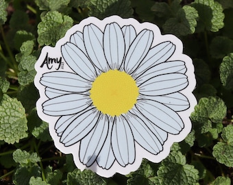 May include: A white daisy sticker with a yellow center and the name "Amy" written in black on the left side of the flower. The sticker is on a bed of green leaves.