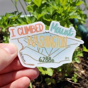 May include: A white sticker with a blue, green, and orange illustration of Mount Washington. The sticker says "I Climbed Mount Washington" and "6288 ft" with the text "Cog Railway" at the bottom left and "Amy" at the bottom right.