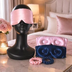 May include: A black mannequin head wearing a light pink silk sleep mask. Below are two additional sleep masks in light pink and navy blue, along with two matching silk scrunchies in light pink and navy blue, all featuring a smooth, shiny finish.
