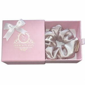 May include: A light pink box with a white bow and the text "HER BEAUTY SUITE" contains a light pink satin scrunchie. The scrunchie is made of a soft, shiny material. The box is open, revealing the scrunchie.