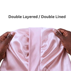 May include: A double-layered, double-lined satin bonnet in a soft pink colour. The bonnet is held open, showcasing the interior lining and the smooth, shiny texture of the fabric. The text "Double Layered / Double Lined" is displayed in black.