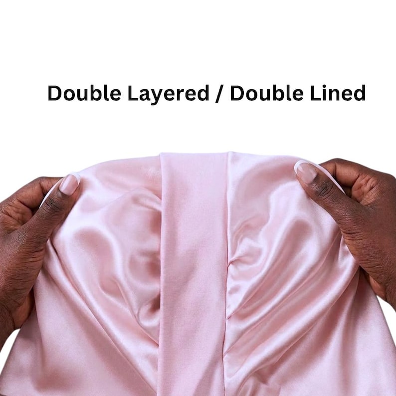 May include: A double-layered, double-lined satin bonnet in a soft pink colour. The bonnet is held open, showcasing the interior lining and the smooth, shiny texture of the fabric. The text "Double Layered / Double Lined" is displayed in black.
