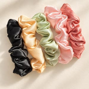 May include: A set of five satin hair scrunchies arranged in a row, featuring colors of black, pale yellow, sage green, light pink, and dusty rose. Each scrunchie has a gathered, ruffled fabric texture with a smooth, shiny finish and elastic interior.