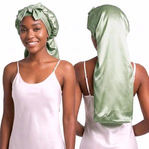 May include: Two images of a person wearing a sage green satin bonnet. The bonnet has a bow on top and a long, flowing tail. The person is wearing a white camisole. The background is white.