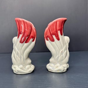 May include: A pair of white ceramic salt and pepper shakers with red painted accents. The shakers are shaped like flames and have a textured surface.