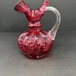 May include: A small, ruby-red glass pitcher with a clear glass handle. The pitcher has a ruffled rim and a bulbous body with a textured, beaded design. The handle is arched and attached to the top and side of the pitcher.