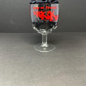 May include: A clear glass goblet with a red design that says "Keep on Truckin'" and features a cartoon image of people riding on a truck. The goblet is sitting on a black surface.