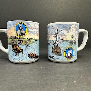 May include: Two white ceramic mugs with blue and yellow designs. One mug features a portrait of a man with the text "The First Group of German Immigrants" and the other mug features a ship with the text "300 Years of German Settlement in America".
