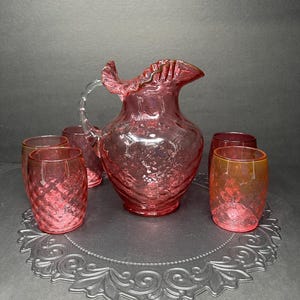 May include: A set of six pink glass tumblers and a pitcher with a ruffled rim. The glasses and pitcher have a textured, bubbly surface.