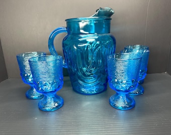 May include: A set of blue glass pitcher and tumblers. The pitcher has a textured surface and a wide mouth. The tumblers have a footed base and a textured surface.