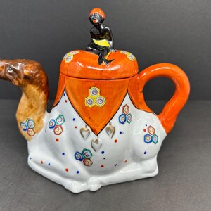 May include: A ceramic teapot shaped like a camel with a small figure of a woman sitting on top. The teapot is decorated with orange, white, and blue geometric patterns.