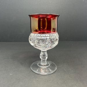 Puede incluir: Una copa de vidrio transparente con una parte superior roja iridiscente. La copa tiene una base con tallo y un patrón decorativo alrededor del cuenco.