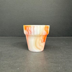 Puede incluir: Un pequeño macetero de vidrio blanco y naranja con un diseño de remolino. El macetero tiene un fondo redondeado y una parte superior ligeramente acampanada.