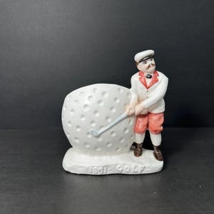May include: White ceramic figurine of a golfer holding a golf club and standing next to a large white golf ball. The base of the figurine is decorated with the words "1891 GOLF".