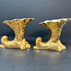 May include: Two gold-plated ceramic vases with a textured surface and a trumpet-like shape. The vases are on a black surface.
