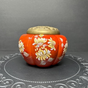May include: A red ceramic jar with a brass lid and white floral designs. The jar is sitting on a black and silver patterned surface.