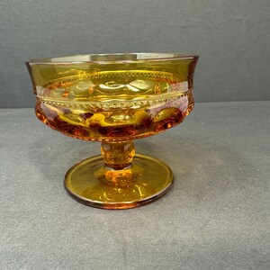 May include: A clear amber glass pedestal bowl with a decorative pattern. The bowl has a wide, round base and a smaller, round top.