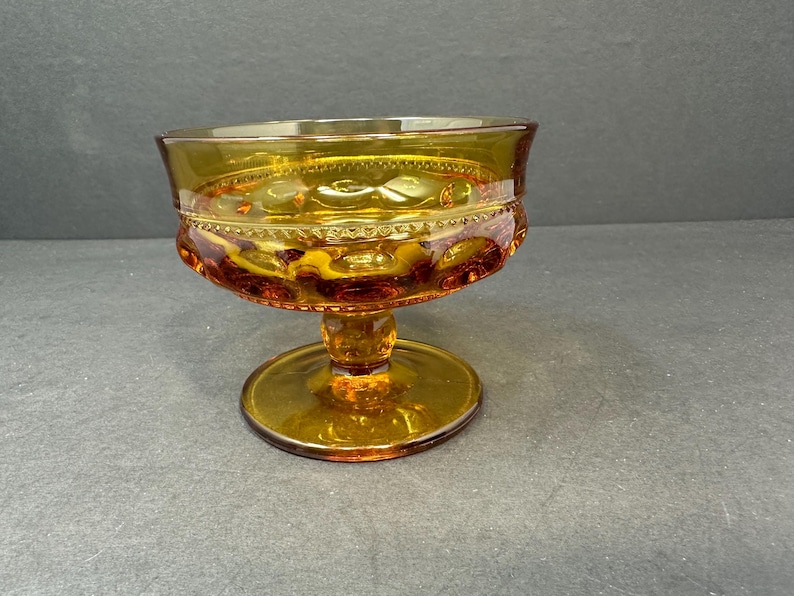 May include: A clear amber glass pedestal bowl with a decorative pattern. The bowl has a wide, round base and a smaller, round top.