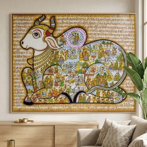 May include: A large, framed artwork featuring a detailed illustration of a cow. The cow is depicted in a seated position, with intricate patterns and figures filling its body. The background is filled with script, and the artwork is framed in gold.