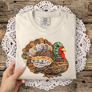 Könnte beinhalten: Ein cremefarbenes Comfort Colors T-Shirt mit einer detaillierten Illustration einer Truthahn. Der Truthahn hat einen grünen Kopf, eine rote Wamme und einen Fächer aus braunen, beigen und grünen Federn. Das T-Shirt ist gefaltet und liegt auf einer weißen Spitzendecke.