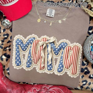 May include: A taupe Comfort Colors t-shirt with a patriotic "MAMA" design. The letters are blue with white stars and red stripes, trimmed with cream lace and a cream bow. A gold necklace and red baseball cap are also visible.