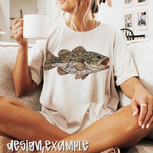 May include: Cream-colored t-shirt featuring a detailed illustration of a fish with open mouth. The fish has a camouflage pattern on its back and a light-colored belly. The words "design example" are printed at the bottom of the image.
