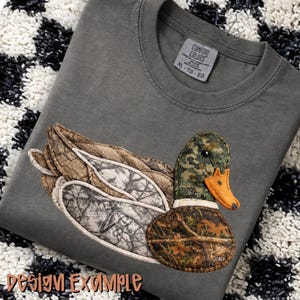 May include: Gray Comfort Colors XL t-shirt with a detailed duck applique. The duck features camouflage patterns in brown, green, and white, with an orange beak. The words "design example" are at the bottom of the shirt.