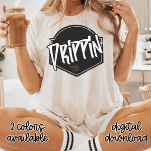 May include: A cream-coloured t-shirt featuring a black and white graphic with the word "DRIPPIN" in a stylized font. The shirt also has the text "2 colours available" and "digital download". A glass jar with a straw is held in the left hand.