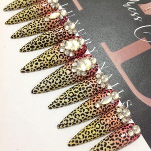 May include: A set of ten stiletto-shaped press-on nails featuring a gradient transition from yellow to orange and red, covered in a black leopard print pattern. Each nail is accented with clear, faceted rhinestones near the cuticle area.