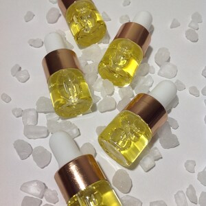 May include: Five small clear glass dropper bottles filled with a vibrant yellow liquid. Each bottle features a metallic copper-colored collar and a white rubber dropper top, arranged among scattered pieces of translucent white rock salt crystals.