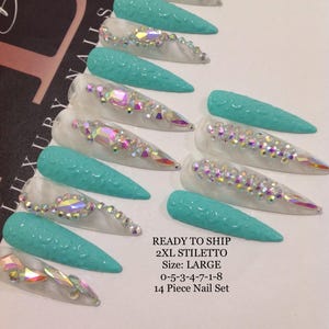 May include: A set of 14 XL stiletto-shaped artificial nails. The nails alternate between a clear base with iridescent rhinestones and a textured turquoise finish. The text "READY TO SHIP" and nail size information are visible.