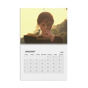 May include: A 2026 wall calendar featuring a scene from a movie. The calendar shows the month of January with a photo of a person reading a book. The calendar has a white background with a spiral binding at the top.