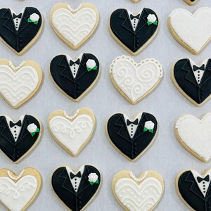 May include: A collection of heart-shaped sugar cookies decorated with royal icing. Some cookies feature black tuxedo designs with white shirts and small white flowers, while others display white wedding dress patterns with intricate piping.