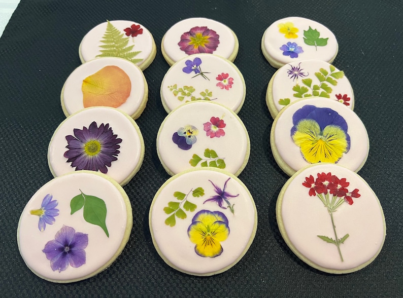 May include: Twelve round sugar cookies decorated with pressed flowers. The cookies are a light pink color and feature a variety of flowers, including purple, yellow, red, and green.