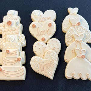 May include: A set of six decorated sugar cookies featuring wedding themes. The cookies are decorated with white icing and rose gold accents. The cookies include a tiered wedding cake, two heart-shaped cookies with the word "love" written on them, and three cookies shaped like wedding dresses.