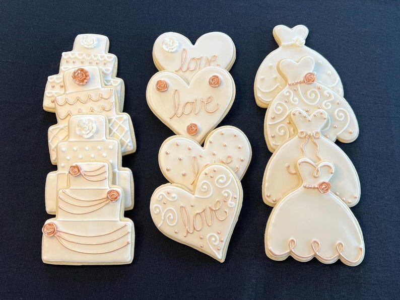 May include: A set of six decorated sugar cookies featuring wedding themes. The cookies are decorated with white icing and rose gold accents. The cookies include a tiered wedding cake, two heart-shaped cookies with the word "love" written on them, and three cookies shaped like wedding dresses.