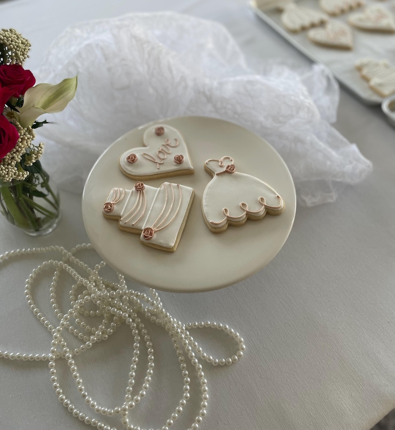 May include: Three white sugar cookies decorated with pink and gold icing. The cookies are shaped like a heart, a wedding dress, and a wedding cake. The heart cookie has the word "love" written on it. The cookies are on a white cake stand.