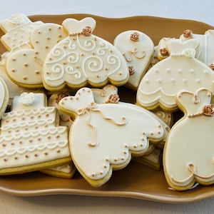 May include: A plate of decorated sugar cookies with a wedding theme. The cookies are decorated with white icing and gold accents. Some of the cookies are shaped like dresses, hearts, and cakes.
