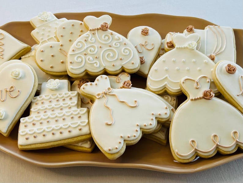 May include: A plate of decorated sugar cookies with a wedding theme. The cookies are decorated with white icing and gold accents. Some of the cookies are shaped like dresses, hearts, and cakes.