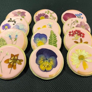 May include: A dozen pink iced cookies decorated with pressed flowers and gold leaf. The flowers include pansies, daisies, and other small blooms.