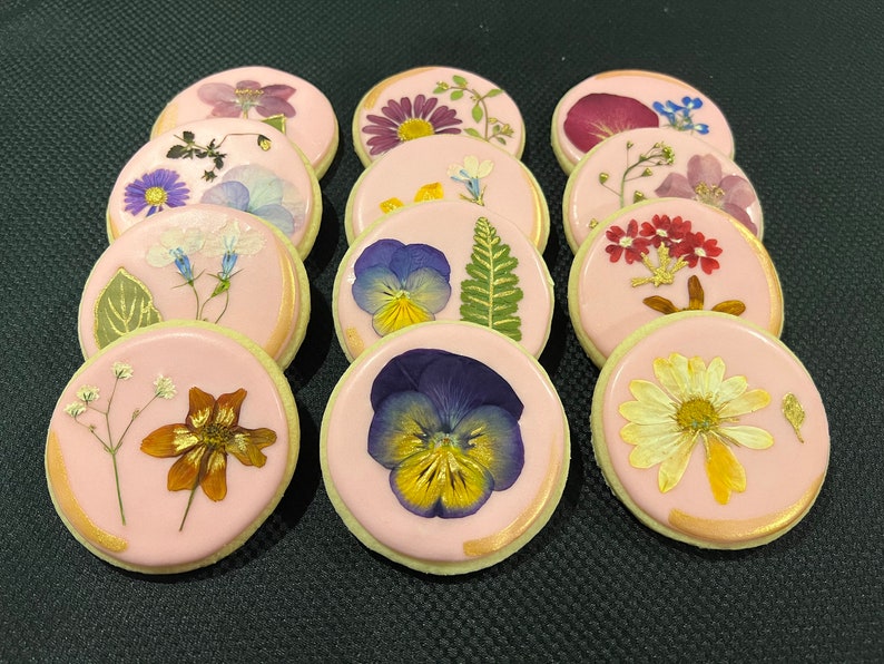 May include: A dozen pink iced cookies decorated with pressed flowers and gold leaf. The flowers include pansies, daisies, and other small blooms.