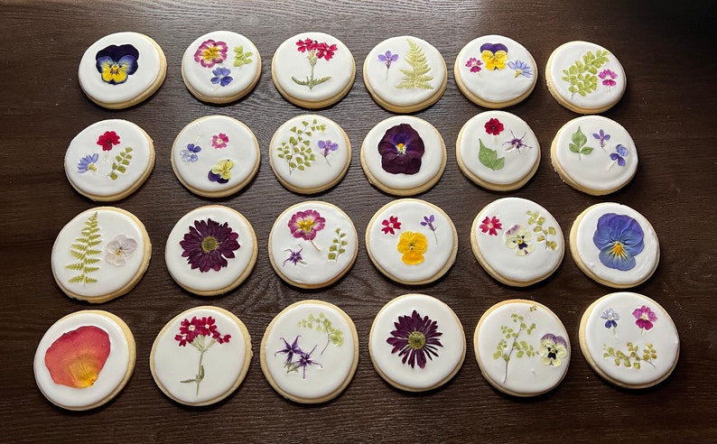 May include: A collection of 20 round sugar cookies decorated with pressed flowers. The cookies are arranged in rows on a wooden surface. The flowers are a variety of colors, including purple, pink, yellow, and green.