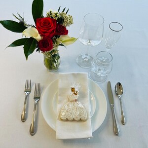 May include: A white ceramic plate with a white and gold iced cookie shaped like a dress. The cookie is on a white napkin. Two silver forks, a silver spoon, and a silver knife are on the table. A bouquet of red roses and white calla lilies is in a clear glass vase.