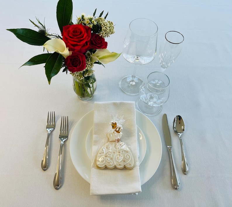 May include: A white ceramic plate with a white and gold iced cookie shaped like a dress. The cookie is on a white napkin. Two silver forks, a silver spoon, and a silver knife are on the table. A bouquet of red roses and white calla lilies is in a clear glass vase.