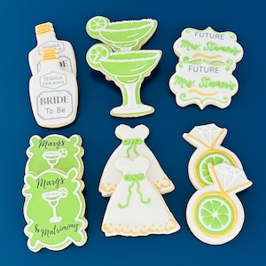 May include: Assortment of decorated sugar cookies on a blue background. The cookies include margarita glasses, a tequila bottle, a wedding dress, diamond rings, and cookies with text that reads "Bride To Be", "Margs & Matrimony", and "Future Mrs. Stanssiy".