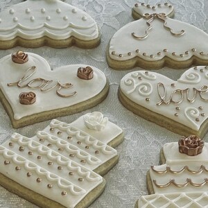 May include: Assortment of white and gold decorated sugar cookies. The cookies are shaped like hearts, dresses, and wedding cakes. Some cookies have the word "love" written on them in gold icing.