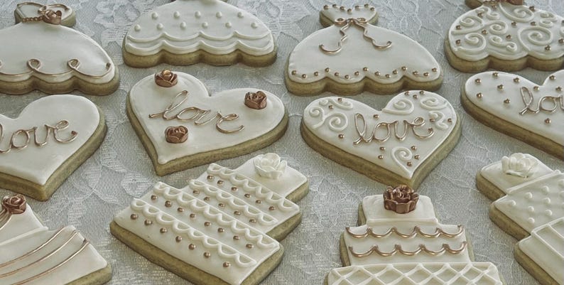 May include: Assortment of white and gold decorated sugar cookies. The cookies are shaped like hearts, dresses, and wedding cakes. Some cookies have the word "love" written on them in gold icing.