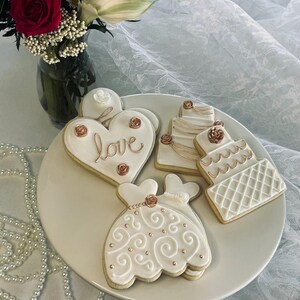 May include: Four decorated sugar cookies on a white plate. The cookies are decorated with white icing and gold accents. One cookie is shaped like a heart and says "love" in gold lettering. Another cookie is shaped like a wedding dress. The other two cookies are shaped like wedding cakes.